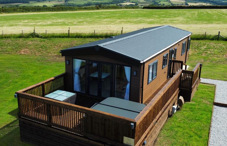 A wooden cabin with a deck and glass doors surrounded by grass and fields at Hazel Lodge in Whitby