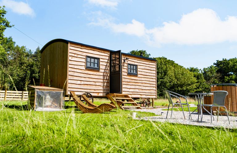 A wooden shepherd hut with wheels on grass near outdoor chairs and a table at Bracken in Broad Langdon near Wainhouse Corner