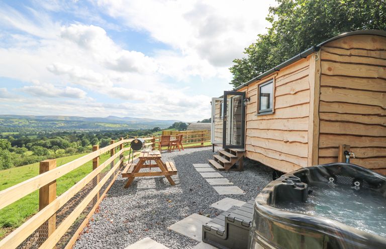 An outdoor area with a wooden cabin a hot tub wooden picnic table chairs and a barbecue with a scenic countryside view at Shepherd's Cwtch in Cilycwm near Llandovery