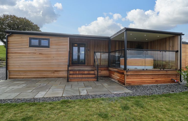 An outdoor view of a wooden cabin with glass doors and a covered patio with a hot tub at Math Lodge 3 Llanelidan near Corwen