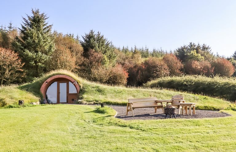 An earth house with a round door surrounded by grass and wooden outdoor furniture in a clearing at Rhosydd in Cyffylliog near Ruthin