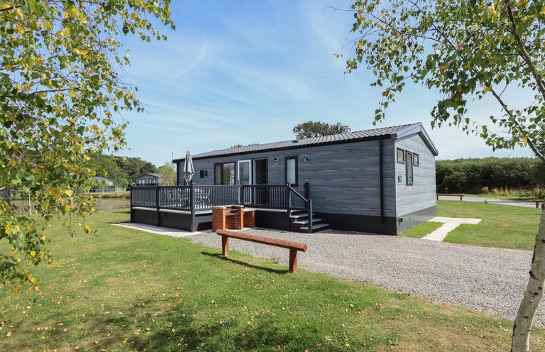 An exterior view of a single-story cabin with a deck and outdoor seating surrounded by grass and trees at Sunset Lodge in Woodhall Spa