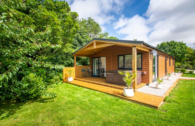 A wooden lodge with a covered porch and outdoor seating surrounded by grass and trees at Maple Lodge in Angarrack