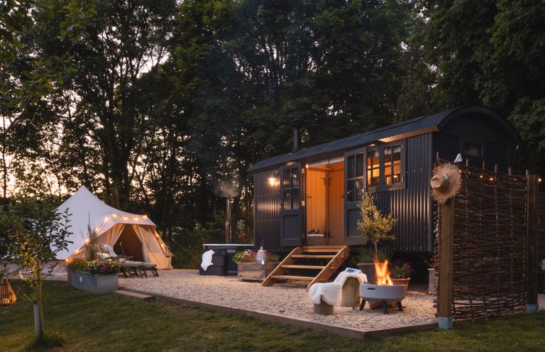 An outdoor camping setup with a tent, a small cabin, lounge chairs, a fire pit, and trees at Moonshine in St Erme near Truro