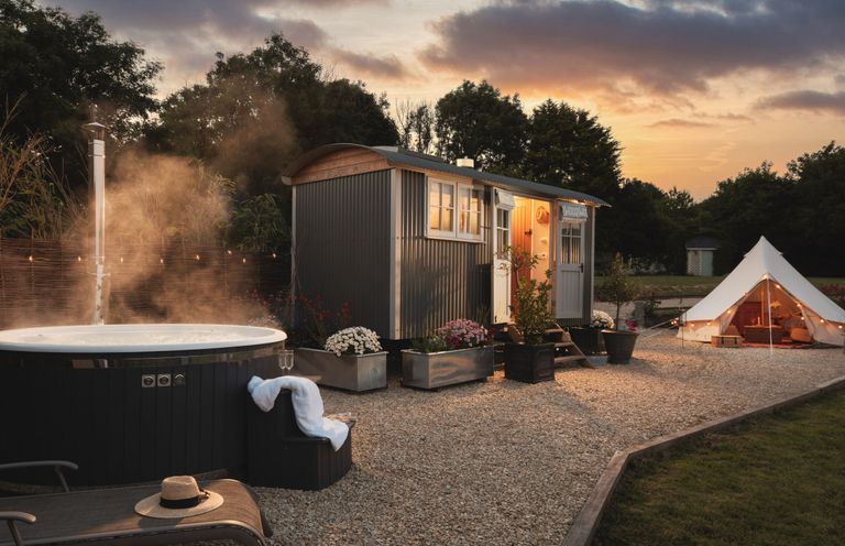 An outdoor area with a hot tub and lounge chair next to a small cabin and a lit tent at sunset at Stargazer in St Erme near Truro