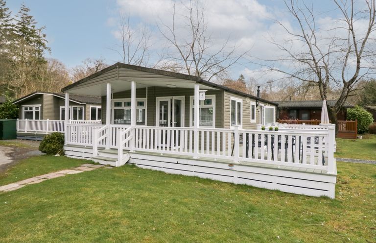 A single-story lodge with a white railing porch on a grassy yard at Cosy Corner in White Cross Bay near Troutbeck Bridge