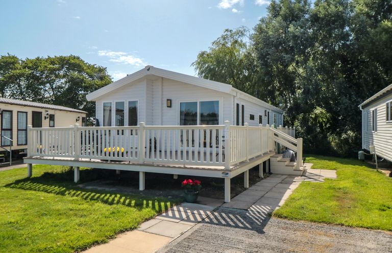 A white mobile home with a fenced deck and steps surrounded by grass and trees at 28 Finch in Cayton Bay