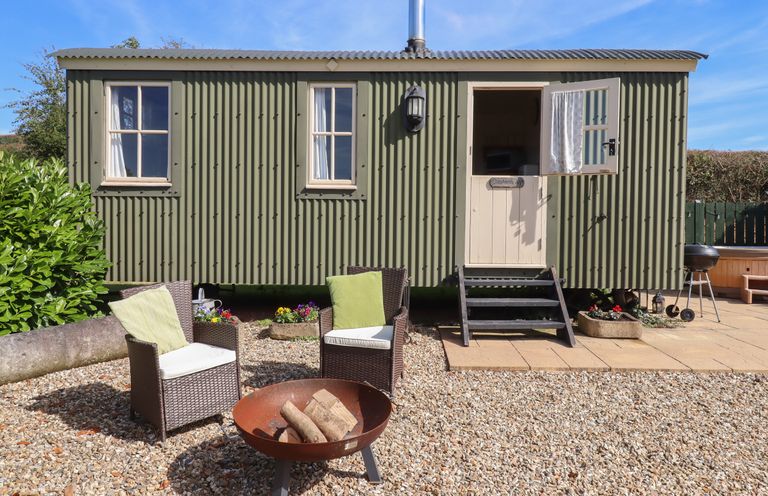 A green metal shepherd hut with two windows and an open door outdoor seating with cushions a fire pit and a barbecue at Shepherds Joy Kennerleigh near Cheriton Fitzpaine