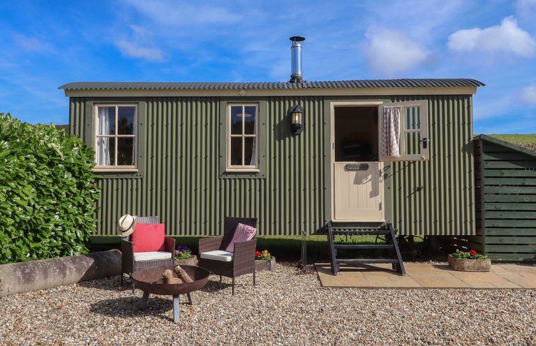 A green corrugated metal cabin with two windows a half door and steps in a gravel yard with two chairs and a fire pit at Shepherds Bliss in Kennerleigh near Cheriton Fitzpaine