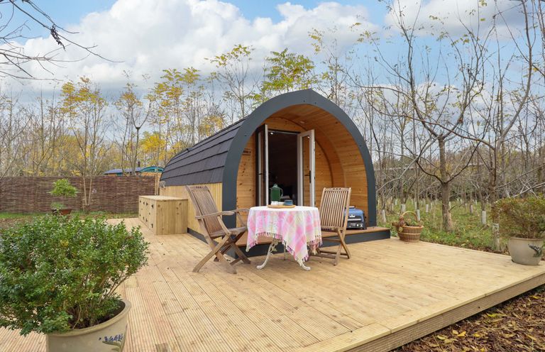 A wooden pod with open double doors on a wooden deck with two chairs and a table with a pink tablecloth surrounded by trees at Hobhole in Keal Cotes near East Kirkby