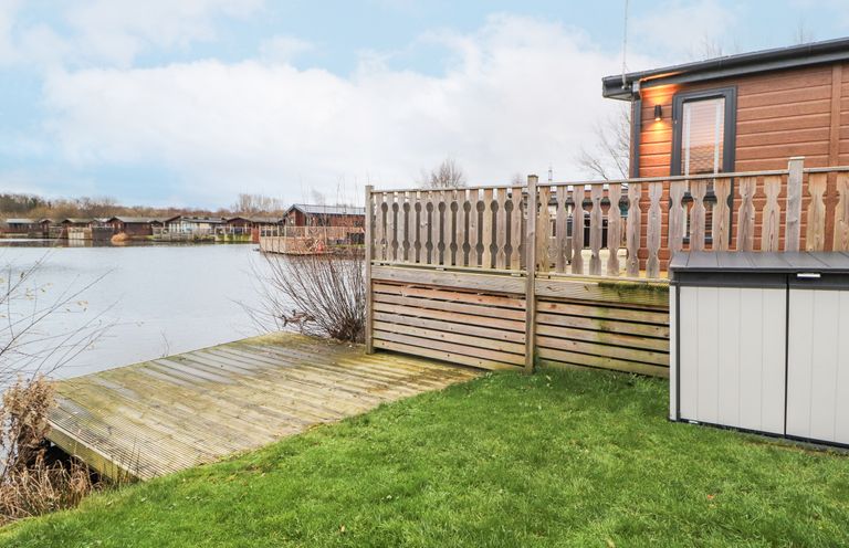 A wooden deck and fenced patio beside a lake with several cabins across the water at 12A Lake Escape in Carnforth