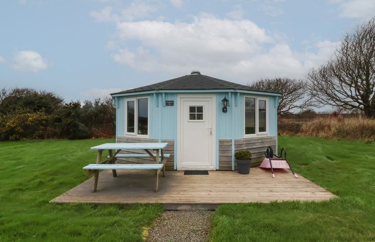 A small blue and wood hexagonal building with white door and windows with picnic table and wheelbarrow on wooden deck at Shipload Bay near Hartland