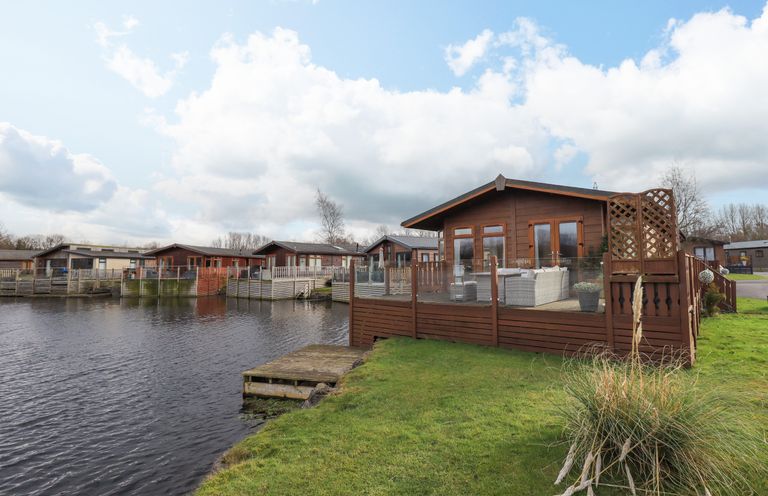 A wooden cabin with a deck and outdoor furniture beside a lake with other similar cabins nearby at Otters Reach near Carnforth
