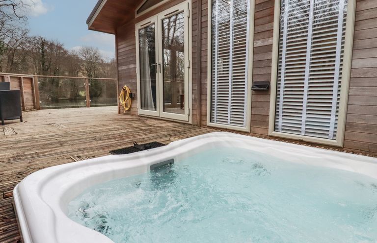 An outdoor deck with a bubbling hot tub near glass doors and windows at 2 Miller's Island Near Lanreath
