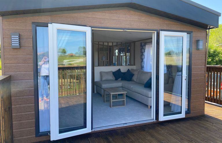 A patio with open glass doors leading to a living room with a grey sofa and wooden coffee table at Maple Lodge in Whitby
