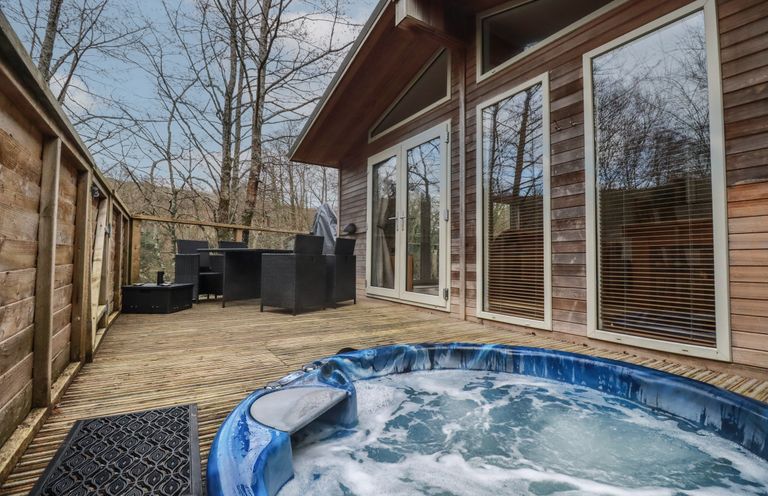 An outdoor deck with a hot tub black chairs and table next to a wooden building with large windows at 1 Miller's Island near Lanreath