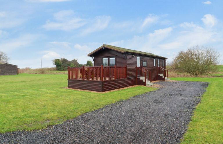 A wooden cabin with a deck and steps surrounded by grass with a gravel driveway at Cuckoo in Ramsey Huntingdon