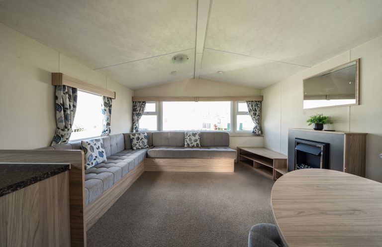 A living room with an L-shaped sofa cushions a round table a fireplace and windows with curtains at Ripples & Reeds - Billing Aquadrome in Billing Aquadrome Holiday Park