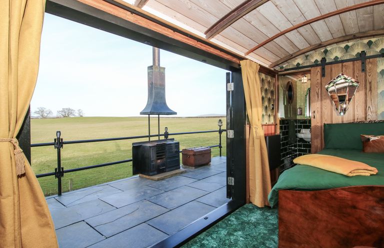 An open room with a bed green carpet and yellow curtains opening to an outdoor patio with a black stove and grassy fields at GWR Wagon - Victoria Station in Ludlow