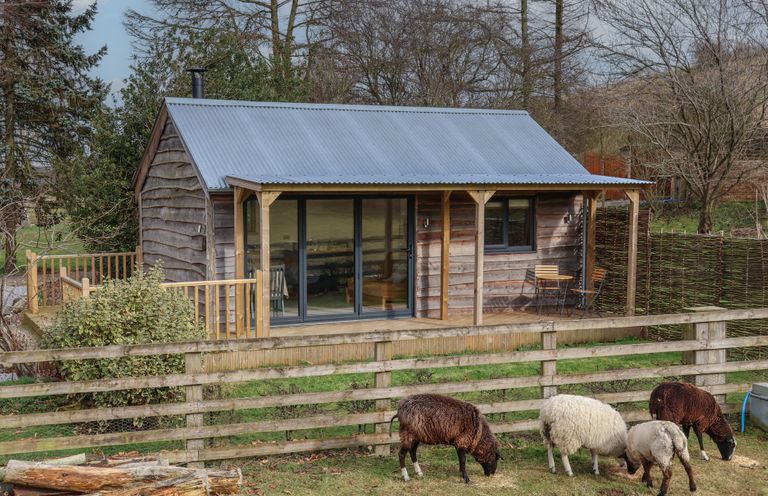 A wooden cabin with a metal roof surrounded by a wooden fence and sheep grazing on grass at Beech Tree Cabin in Goodmanham near Market Weighton