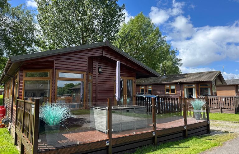 A wooden cabin with glass railing and outdoor seating on a deck surrounded by trees at Lakeside 18 in Carnforth