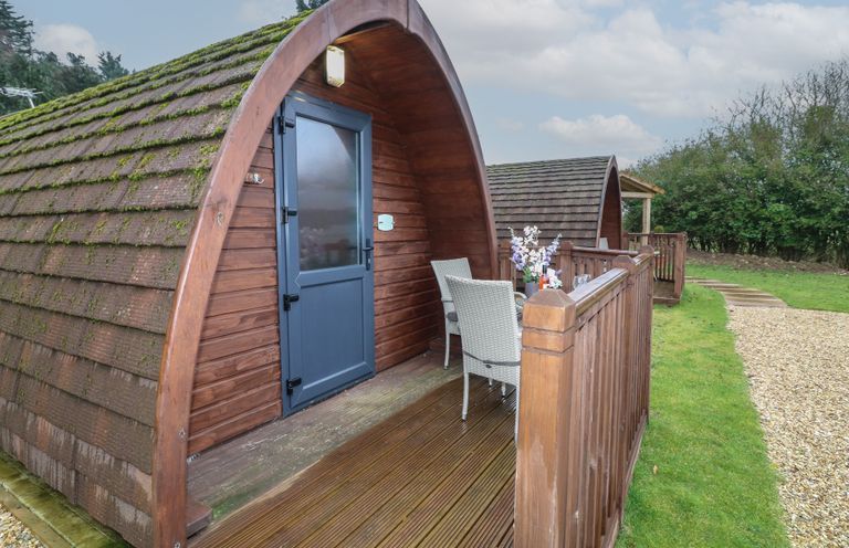 Two wooden pods with blue entrance doors and outdoor seating on a wooden deck near a gravel path and grassy area at Chantelles in Holbeach