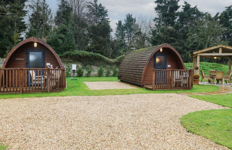 Two wooden camping pods with small porches on grass next to a gravel path and a wooden gazebo with table and chairs at Charlottes in Holbeach