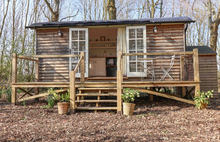 A wooden shepherds hut with open double doors and a small porch with steps and a metal table and chairs at Woodlands Retreat Shepherds Hut Brundish near Laxfield
