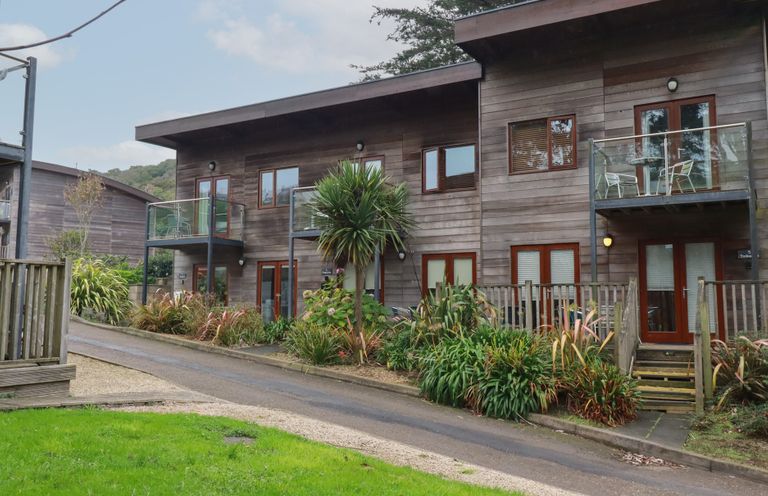 A wooden two story building with balconies and glass railings surrounded by plants and a road at Treburgett in Porthtowan
