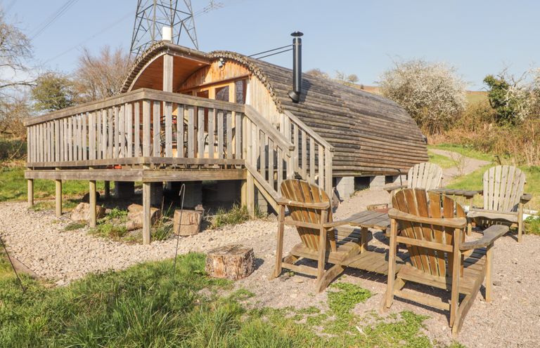 A wooden cabin with a deck and outdoor chairs on gravel surrounded by grass at Cabanau Bach in Bedwas