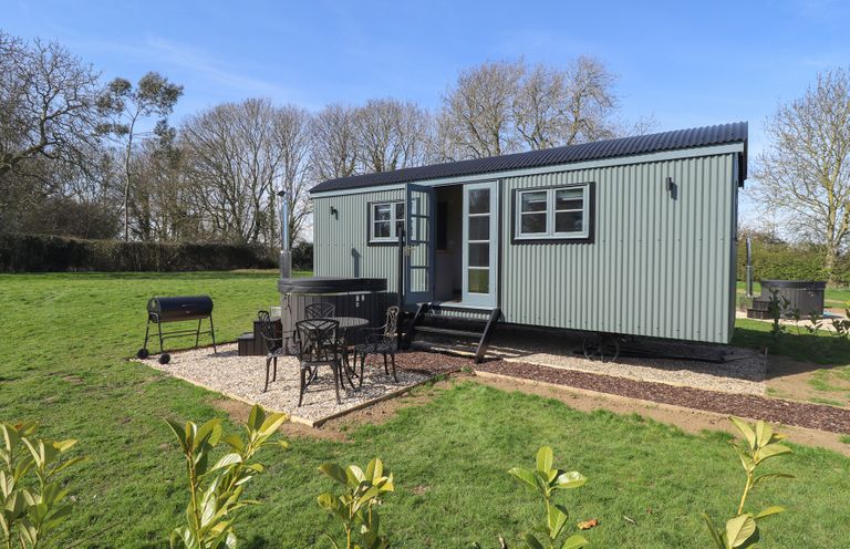 A green metal shepherd hut with a small outdoor seating area and a barbecue on grass in a rural setting at Buttercup in Ashby-cum-Fenby near Waltham