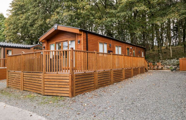 A wooden cabin with a fenced deck and gravel driveway surrounded by trees at Water's Reach White Cross Bay Holiday Park near Troutbeck Bridge