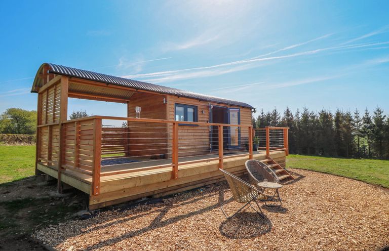 A wooden shepherd hut with a covered porch and two chairs on wood chip ground at Fir Tree Shepherd's Hut in Llanon