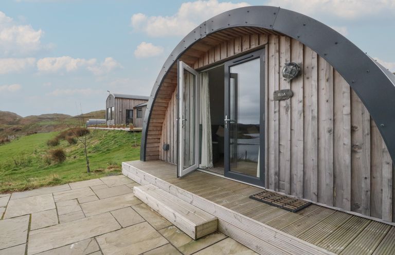 An arched wooden cabin with glass doors open to a stone patio and grassy landscape at Sula Cullipool Isle Of Luing