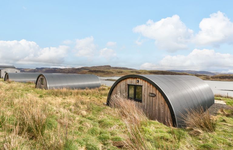 A row of curved metal and wooden cabins near water with hills in the background at Scarba in Cullipool Isle Of Luing