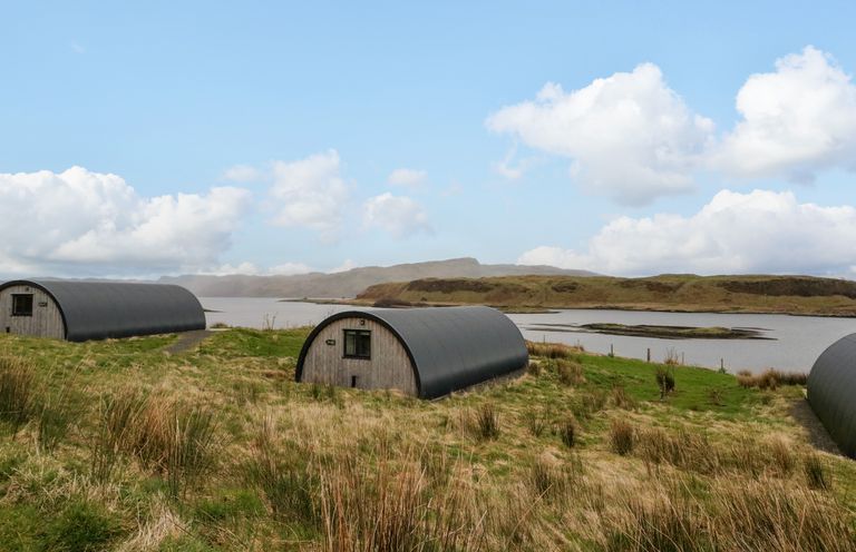 Three curved black roof huts on grassy land by water with low hills in the background at Fladda Cullipool Isle Of Luing