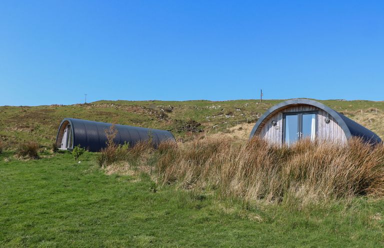 Two curved metal and wooden structures on grassy land with hills and clear blue sky at Losal Cullipool Isle Of Luing