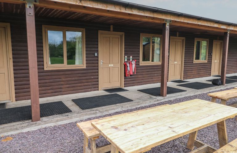 An outdoor seating area with wooden picnic tables in front of a row of wooden cabins with doors and windows at Alder in Cowbridge