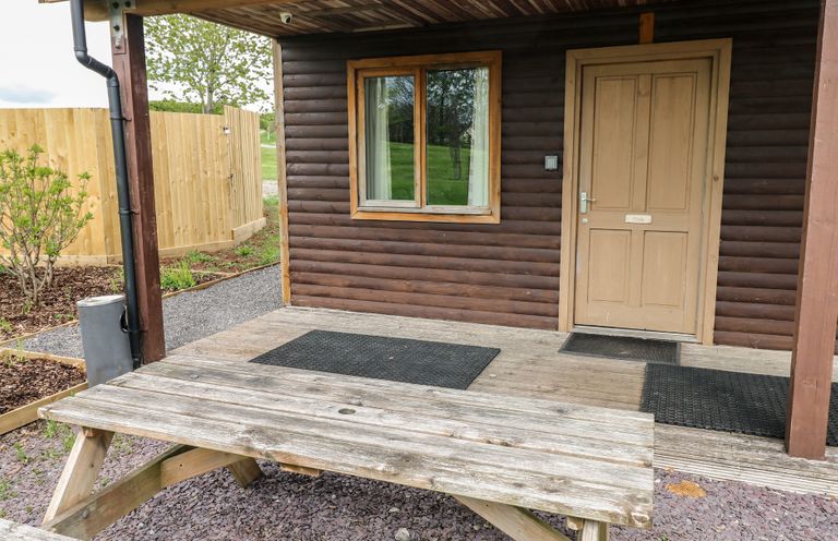 An outdoor wooden picnic table in front of a cabin porch with a window and a door labeled Oak at Oak in Cowbridge