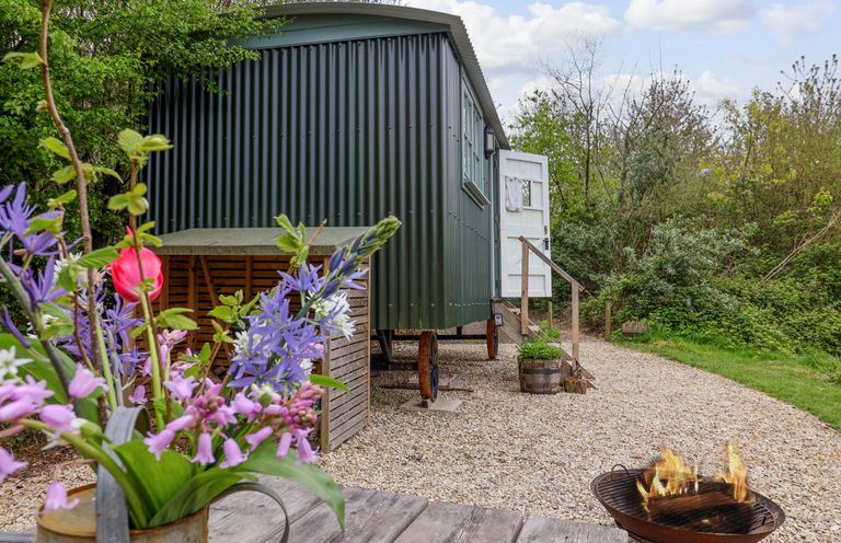 A green shepherd hut with an open white door on a gravel path surrounded by greenery with a fire pit and a wooden table with flowers at Oak Shepherd Hut in Monkton Wyld near Charmouth