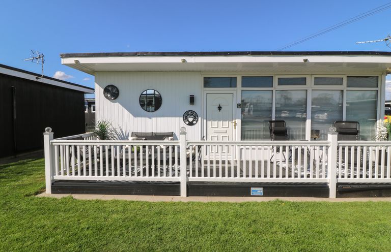 Front view of a white chalet with a fenced deck and outdoor chairs at 56 Cherry Park Chalet in Chapel St Leonards