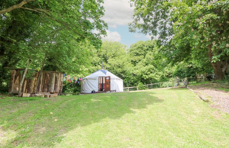 A white yurt with an open door on a grassy hill surrounded by trees and a wooden structure with colorful bunting at Moonlight in Ventnor