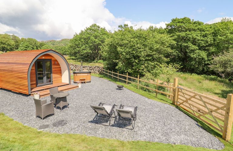 An outdoor area with a wooden pod cabin two chairs and a small table a hot tub and a fenced grassy area at Eirlys in Bontddu near Dolgellau