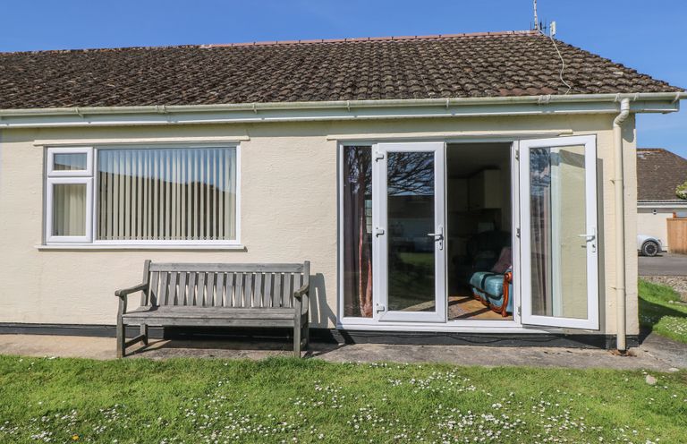 The exterior of a single-story house with a wooden bench on a concrete path and open double glass doors showing a blue armchair inside at Kites - 52 Gower Holiday Village in Scurlage near Port Eynon and Rhossili