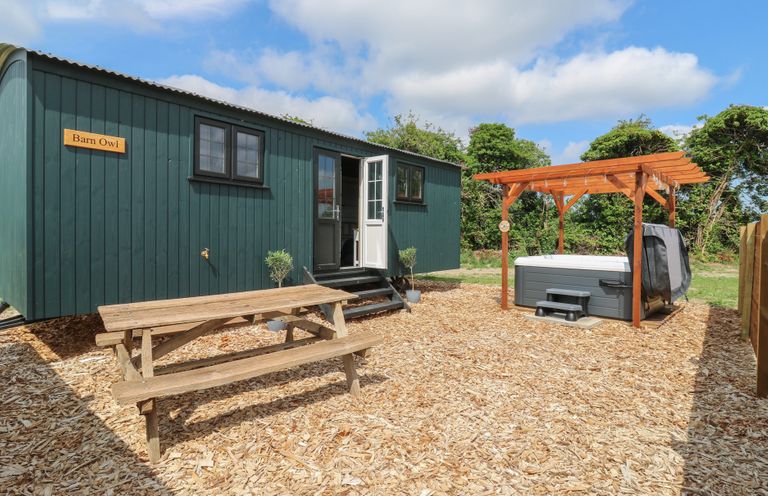 An outdoor area with a green wooden cabin labeled Barn Owl a wooden picnic table and a hot tub under a wooden pergola at Barn Owl in Lode near Bottisham