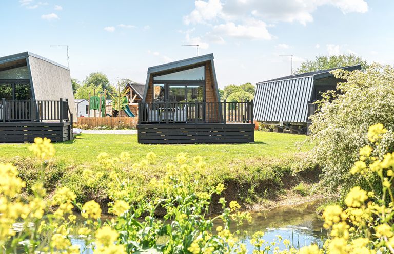 A grassy area with modern small cabins next to a small pond with yellow flowers and a playground in the background at Celestial Skylight - Billing Aquadrome in Billing Aquadrome Holiday Park