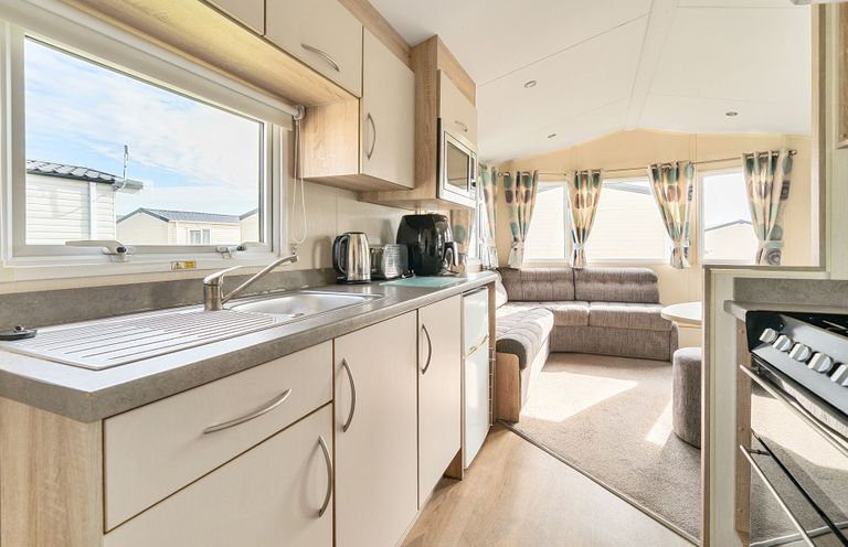 A kitchen area with cupboards sink kettle toaster and microwave leading to a living area with sofa windows and curtains at Golden Dune - Hayling Island in Hayling Island Holiday Park