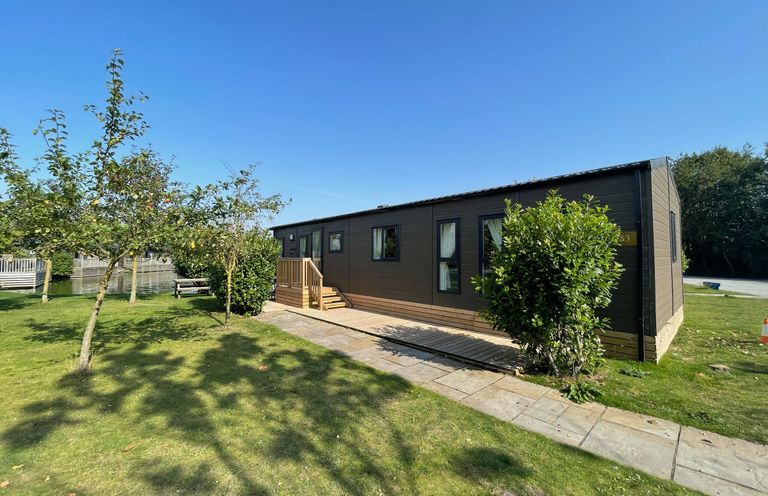 A mobile home with a deck and trees at Acacia Waterside Spa Carlton Miniott near Thirsk