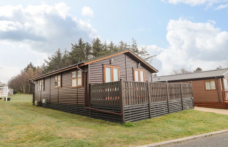 A log cabin with a deck surrounded by grass and trees at Nevaeh Lodge in Longframlington
