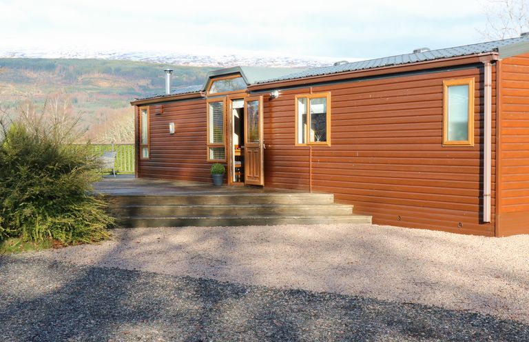 A brown wooden cabin with steps leading to an open door surrounded by gravel and bushes at The Four Oaks Chalet in Blaich near Fort William